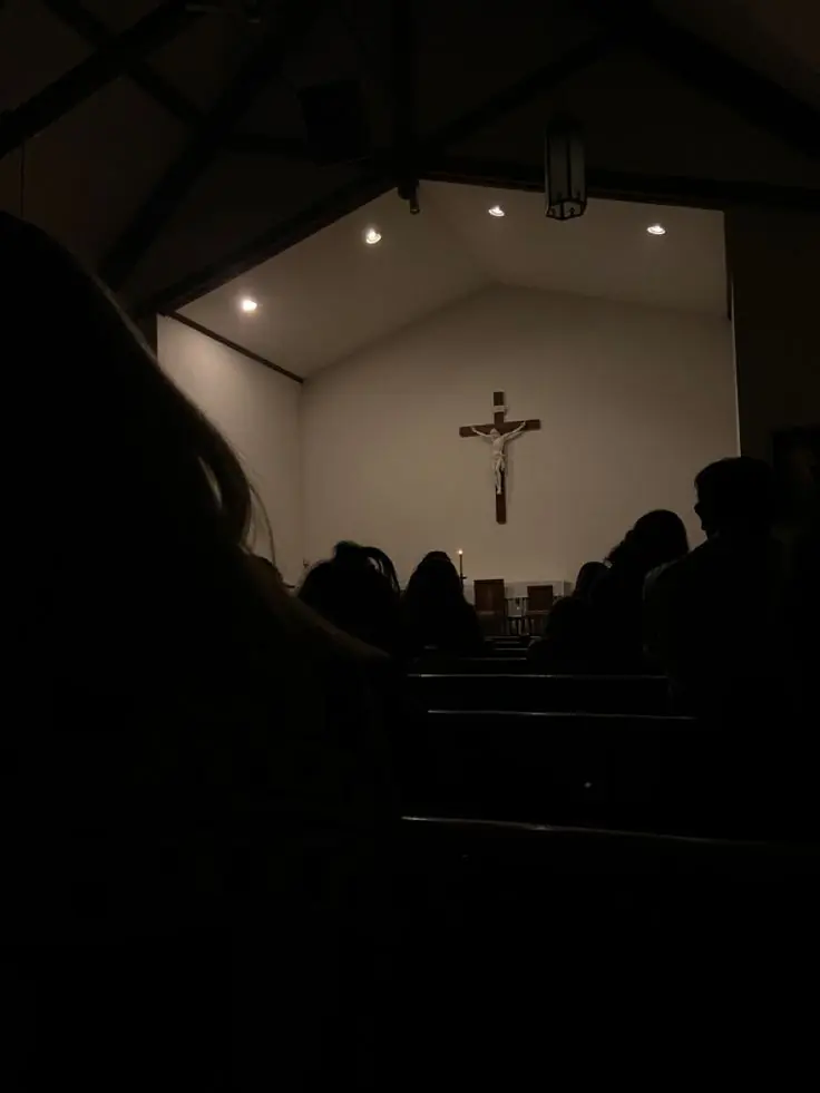 A small, dim church interior. A plain wooden crucifix is lit on a white wall above a low table, with a handful of people seated in the dark below it.